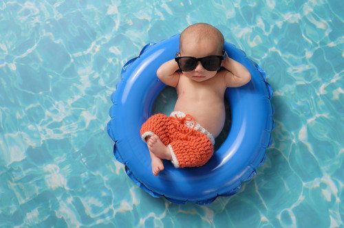 Four week old newborn baby boy sleeping on a tiny inflatable swim ring. He is wearing crocheted board shorts and black sunglasses.